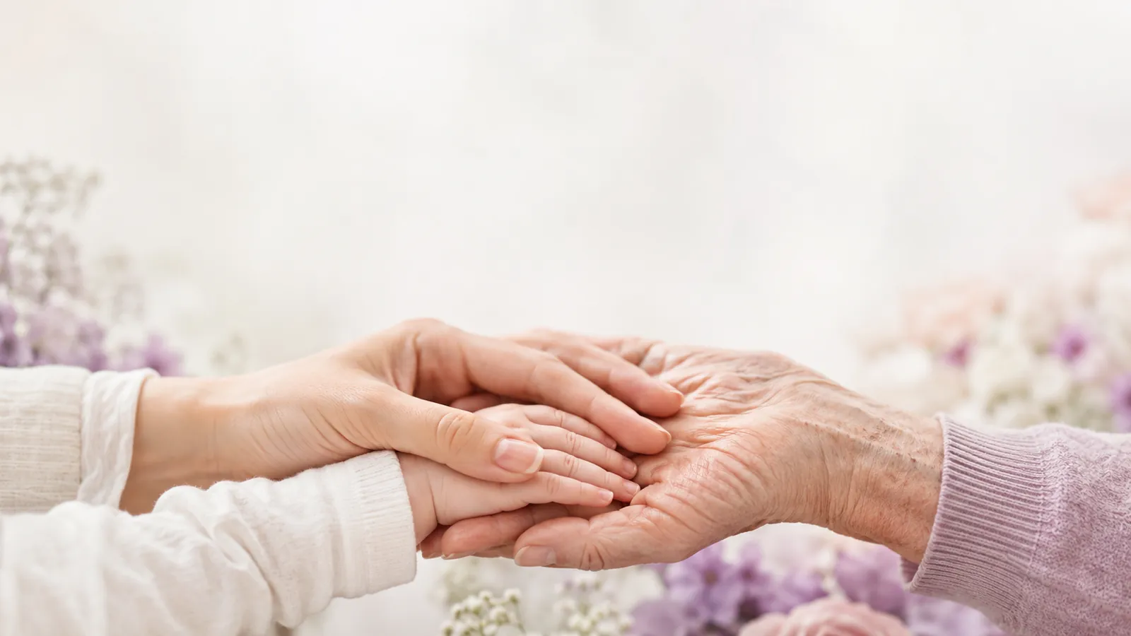 A younger adult's hands gently holding an elder's hand among soft flowers